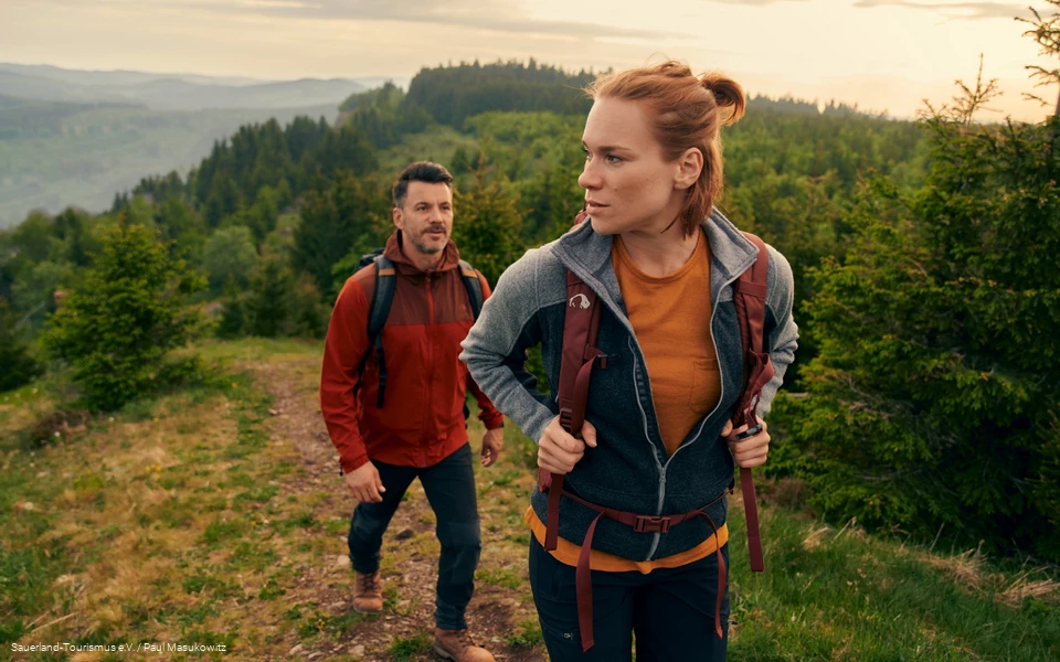 Ein junges Paar wandert einen Berg hinauf. Im Hintergrund ist die üppige, grüne Landschaft zu sehen.